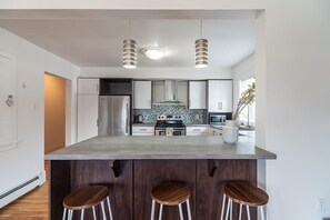 Une cuisine moderne dotée d’un îlot central, d’appareils électroménagers en acier inoxydable et de luminaires suspendus.