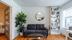 Een moderne woonkamer met een donkere bank, een glazen salontafel, een grote potplant, een boekenkast en een spiegel aan de muur.
