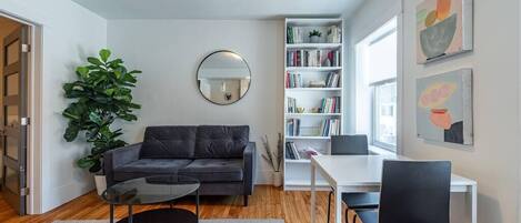 A modern living room with a sofa, coffee table, bookshelf, and a dining area with chairs.