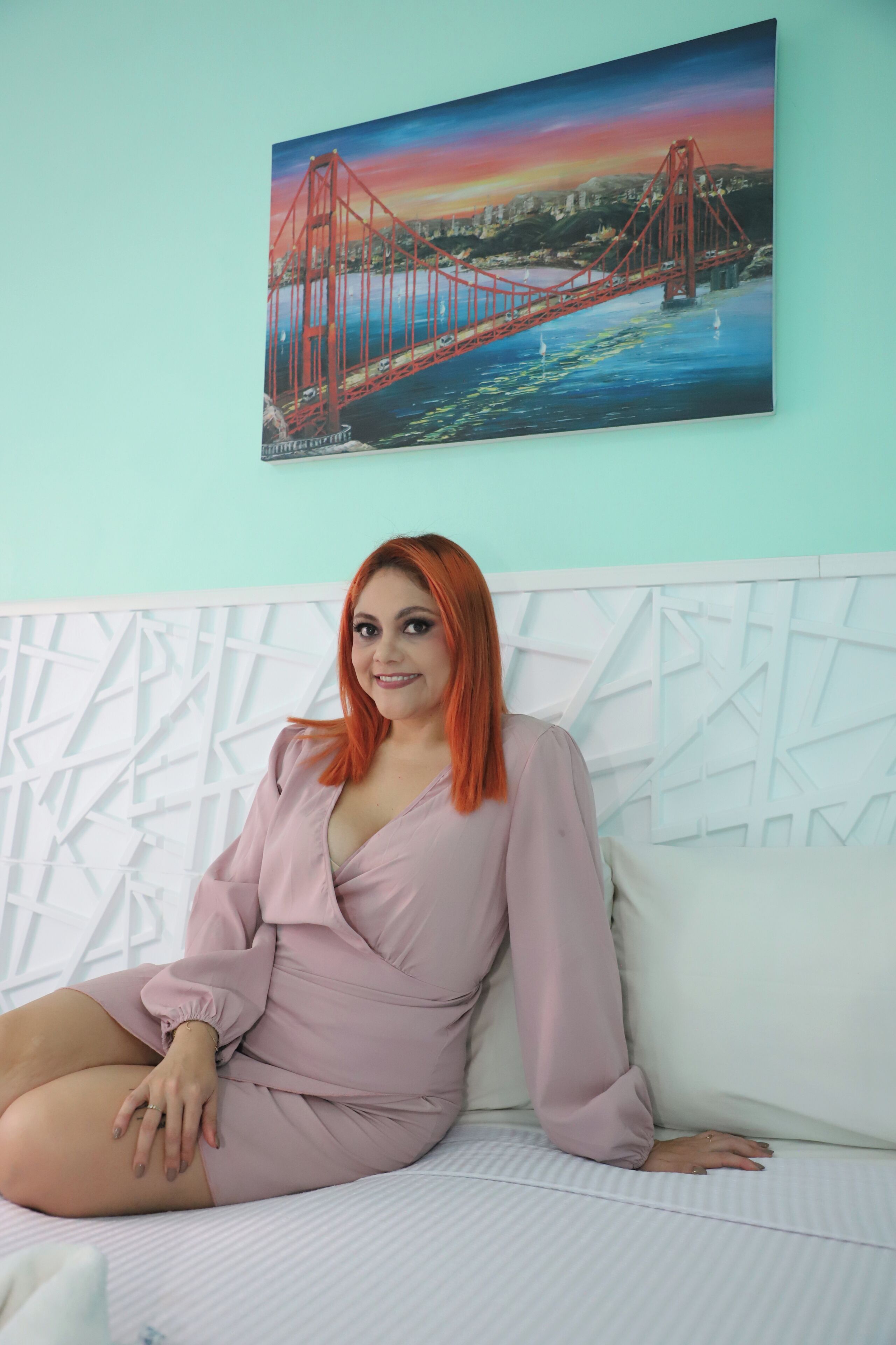 Uma pessoa sentada em uma cama, com uma pintura de uma ponte ao fundo.