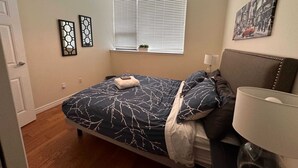 A bedroom with a bed, a bedside table, a lamp, a window with blinds, and a wall-mounted picture. - Family Friendly Downtown Loft with AC (Halifax)
