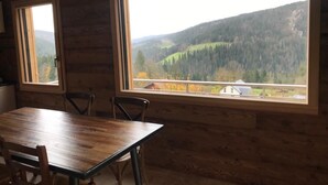 Een houten interieur met een eettafel en stoelen, grote ramen met uitzicht op een bebost gebergte.
