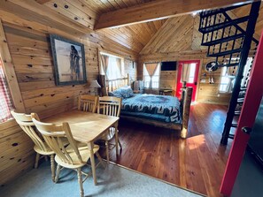 Una cabaña acogedora con interior de madera, una cama con colcha, una mesa de comedor con sillas y una puerta roja.