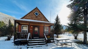 A wooden cabin with a front porch, a bench, and a picnic table, surrounded by snow and trees. - Lemhi Shoshone Cabin, Greyhouse Vacation Rentals (Salmon)