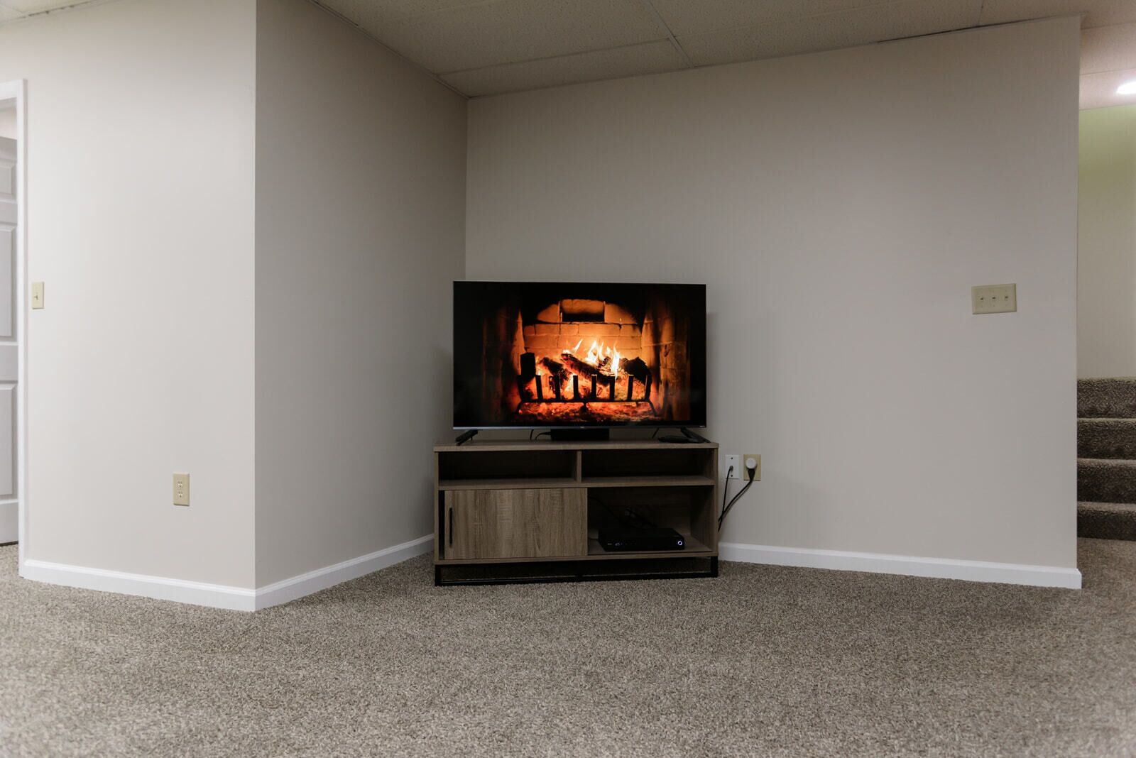 A corner room with a flat-screen TV on a wooden stand, carpeted floor, and white walls.