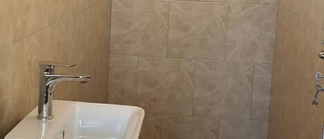 Une salle de bain avec un lavabo blanc, une douche équipée d’un pommeau de douche à main et des murs recouverts de carreaux beiges.