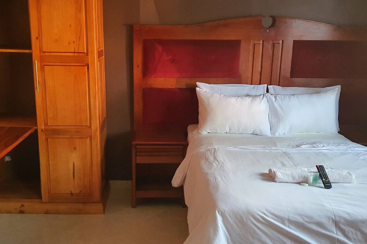 A hotel room with a wooden wardrobe, a bed with white bedding, and a remote control on the bed.