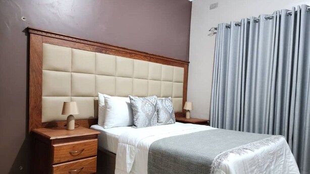 A neatly made bed with a wooden headboard, two bedside tables with lamps, and a grey curtain. - The Princess Guest House (Harare)