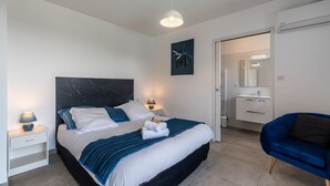 Un dormitorio moderno con una cama grande, mesitas de noche con lámparas, una silla azul y un baño visible a través de una puerta abierta.