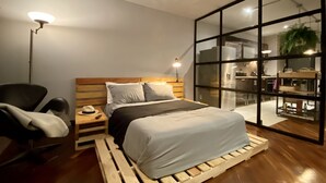 Ein modernes Schlafzimmer mit einem hölzernen Kopfteil, einem Bett mit grauer Bettwäsche, einem Nachttisch mit Lampe, einem Sessel und einem großen Fenster mit Blick auf die Küche.