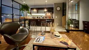 Ein modernes Wohnzimmer mit großem Fenster, einem schwarzen Ledersessel, einem Holztisch mit Zeitschriften und einer Küche im Hintergrund.