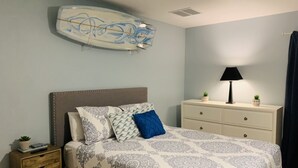 A bedroom with a bed, bedside table, dresser, lamp, and a surfboard on the wall. - Beachfront property with pool view-Aloha (Melbourne Beach)