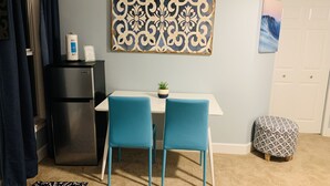A compact living space with a kitchenette, a dining area with two chairs, a patterned wall hanging, and a small potted plant. - Beachfront property with pool view-Aloha (Melbourne Beach)
