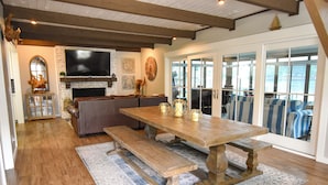 Amplia sala de estar con una mesa de madera, bancos, chimenea y un gran ventanal con vista a un cuerpo de agua.