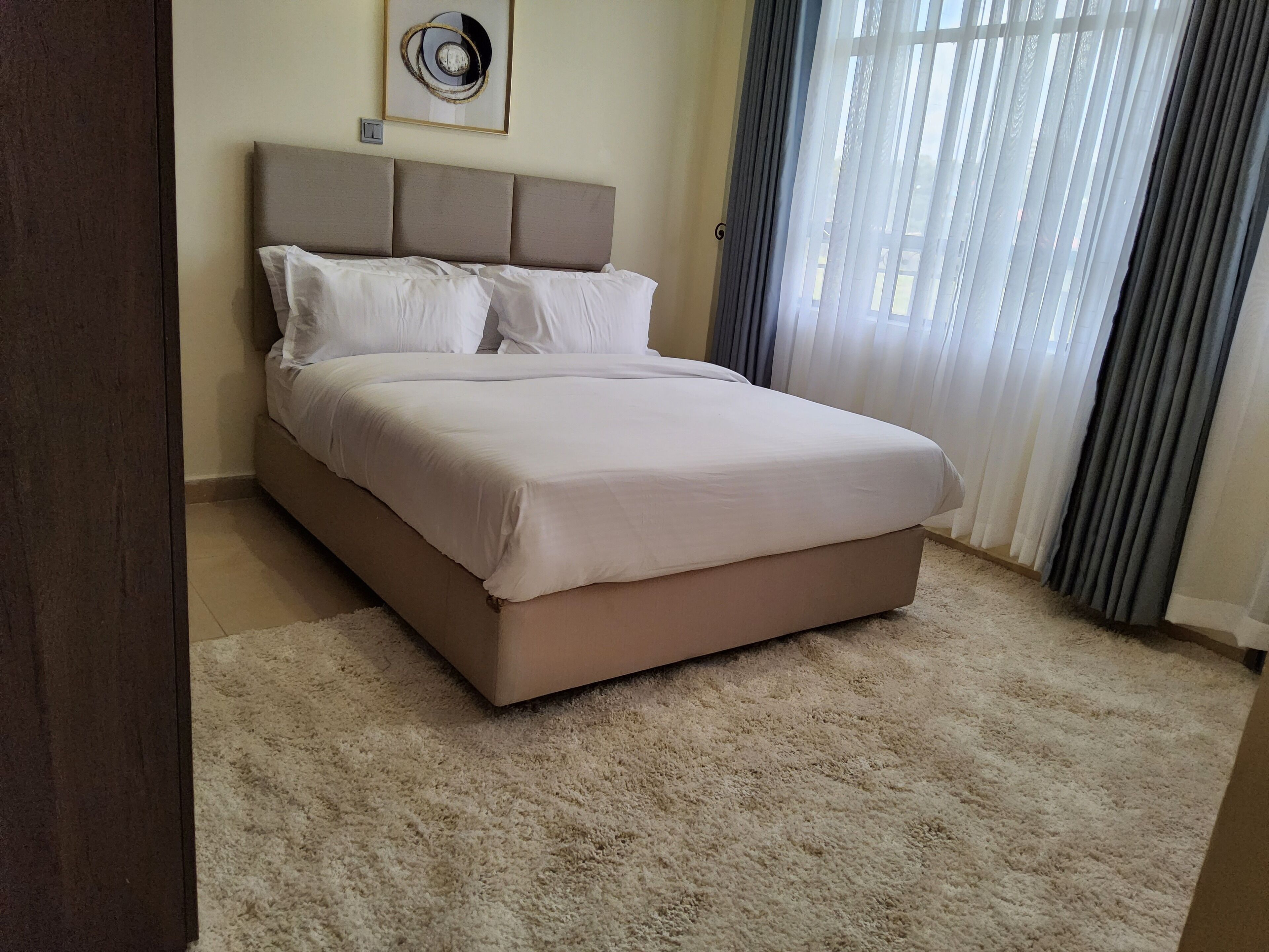 A bedroom with a bed, a headboard, a framed picture on the wall, and a window with curtains.