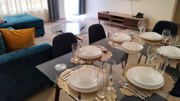 Zona pranzo con tavolo in vetro apparecchiato per sei persone, circondata da divani blu e un televisore che mostra un caminetto.
