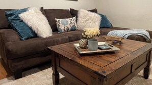 Un salón acogedor con un sofá marrón, cojines decorativos, una mesa de centro de madera con un jarrón de flores y un cuadro en la pared.