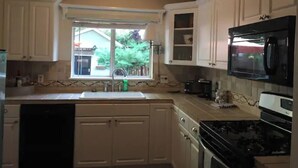 Una cocina con armarios blancos, un microondas negro y una ventana que ofrece vistas a una casa y árboles.