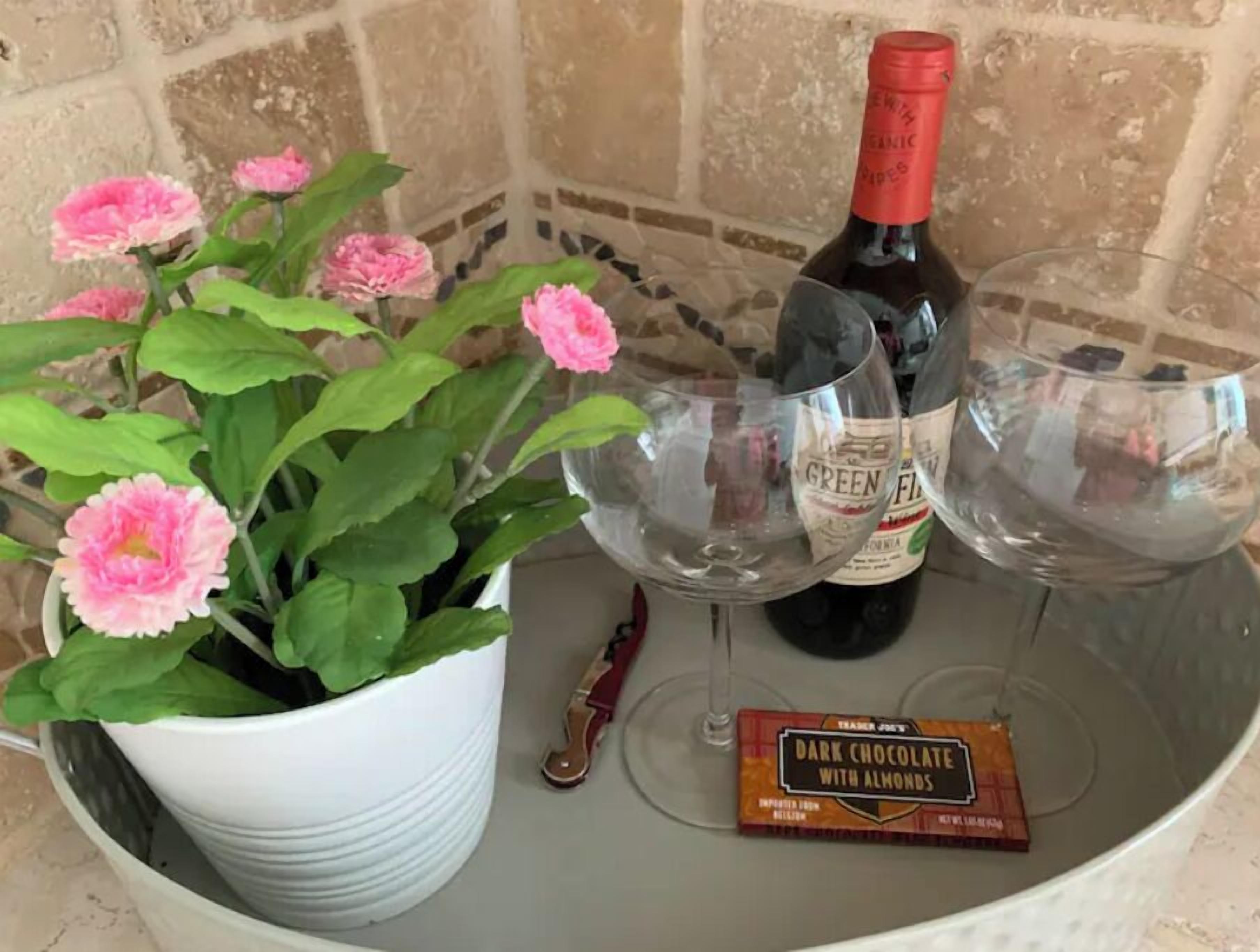 Un pot blanc orné de fleurs roses, deux verres à vin, une bouteille de vin et une tablette de chocolat.