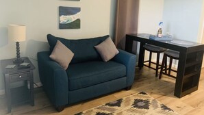 A living room with a blue sofa, two cushions, a small table with a lamp, and a dining area with a table and two chairs. - Cozy Beach Escape -saltwater pool - Poolside Suite (Melbourne Beach)