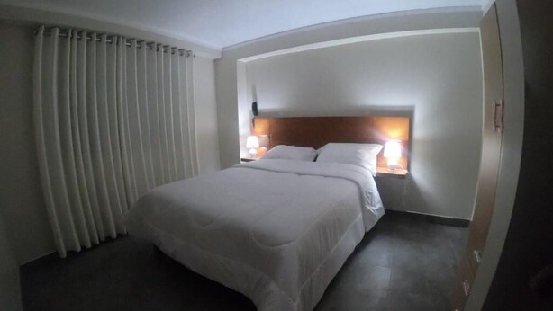 Habitación de hotel con cama, cabecera, dos mesitas de noche con lámparas y una ventana con cortinas.
