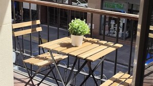 A balcony with a small table and chairs, a potted plant, and a view of a street with a store named 'HERGU.' - Spacious and cosy in a central neighbourhood, Indautxu. (Bilbao)