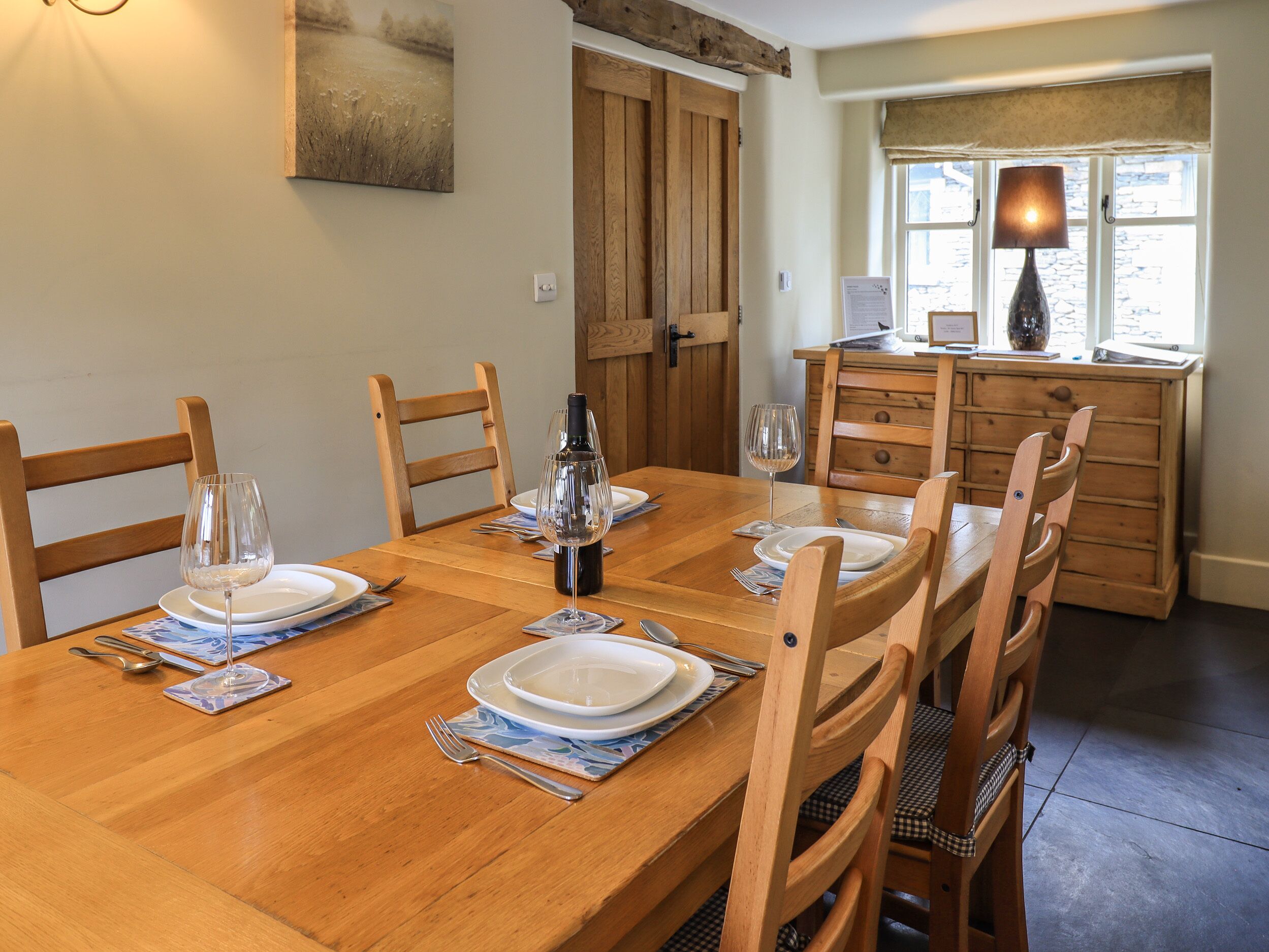 A dining area with a wooden table set for six, including plates, glasses, and cutlery. There are wooden chairs around the table, and a wooden cabinet with drawers is visible in the background.