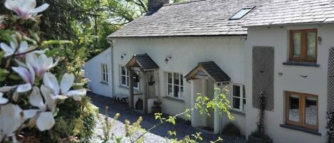 A white cottage with a sloped roof, a small porch, and a garden area with flowers and plants.