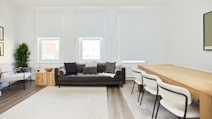 A modern living room with a sofa, a wooden dining table, and chairs. There are windows with blinds, a plant, and a framed picture on the wall.