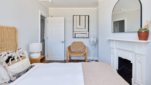 A bedroom with a bed, a chair, a bedside table with a lamp, a white door, a wall-mounted mirror, and a fireplace.