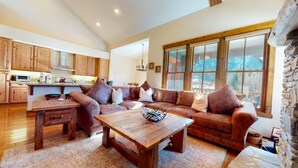 A spacious living area with a wooden coffee table, leather sofa, and a large window offering a view of the outdoors. - Snowcreek Vi The Lodges #1167 4 Bedroom Condo by RedAwning (Mammoth Lakes)