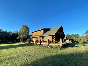 A log cabin with a deck, surrounded by a grassy area and trees. - Handcrafted Log Cabin | Hot Tub | Sauna | GameRoom (Ironwood Township)