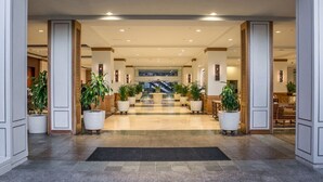 Un vestíbulo de hotel con una escalera central, plantas en macetas y piso de baldosas.