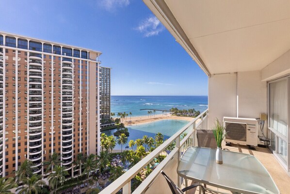 A balcony with a view of a beach and ocean, a pool, and a tall building in the background.