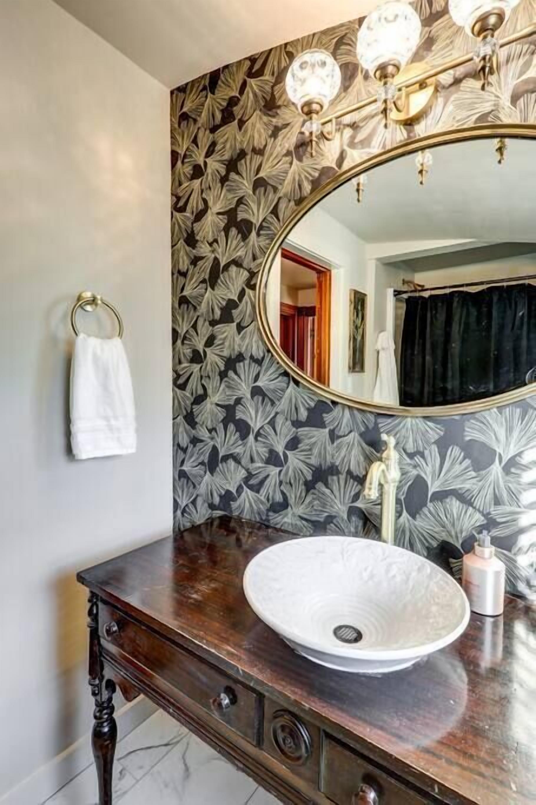 A bathroom with a large oval sink, a wooden vanity, a mirror with a gold frame, and a wall with a leaf-pattern wallpaper.