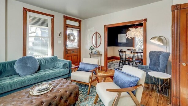 Een woonkamer met een blauwgroene bank, een bruin poef en een houten salontafel. Er is een eethoek met een tafel en stoelen.
