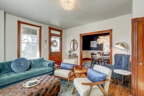 A living room with a teal sofa, a brown ottoman, and a wooden coffee table. There is a dining area with a table and chairs. - The Conservatory-20 min to Lanc! (Mount Joy)