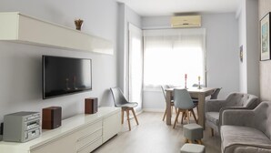 En moderne stue med et fladskærms-tv, et hvidt tv-møbel, grå polstrede lænestole, en grå sofa og et spiseområde med bord og stole.