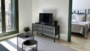 Une chambre d’hôtel moderne avec un téléviseur posé sur une console noire, une petite plante en pot et une vue sur la ville à travers une grande fenêtre.