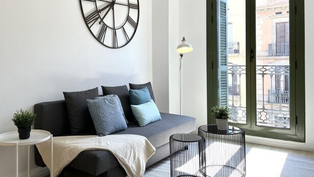 Un salon moderne avec un canapé gris, des coussins à motifs, une grande horloge murale, un lampadaire et une vue sur un balcon donnant sur des immeubles.