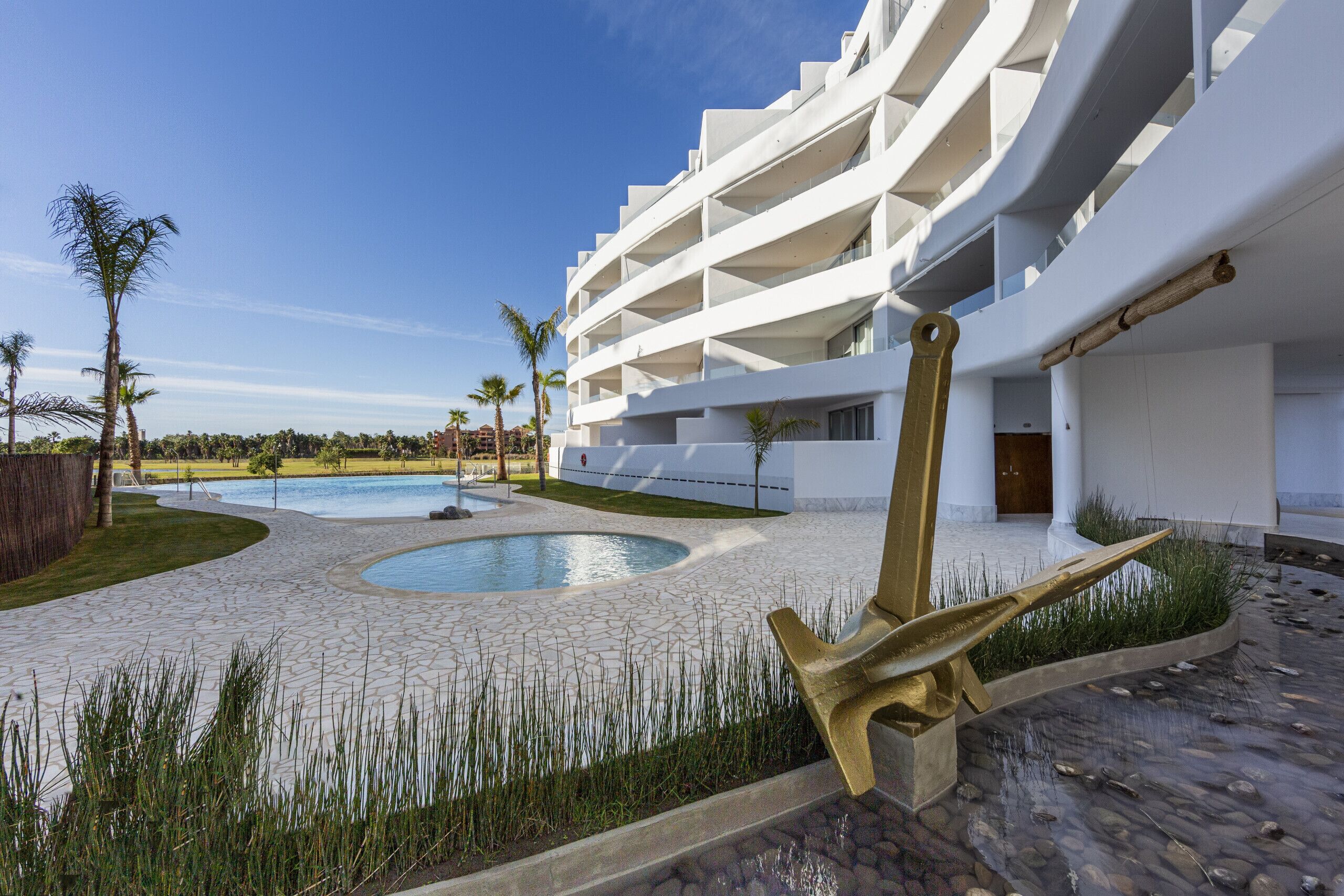 Et moderne hotel med pool, udendørs siddepladser og en markant skulptur af et gyldent anker.