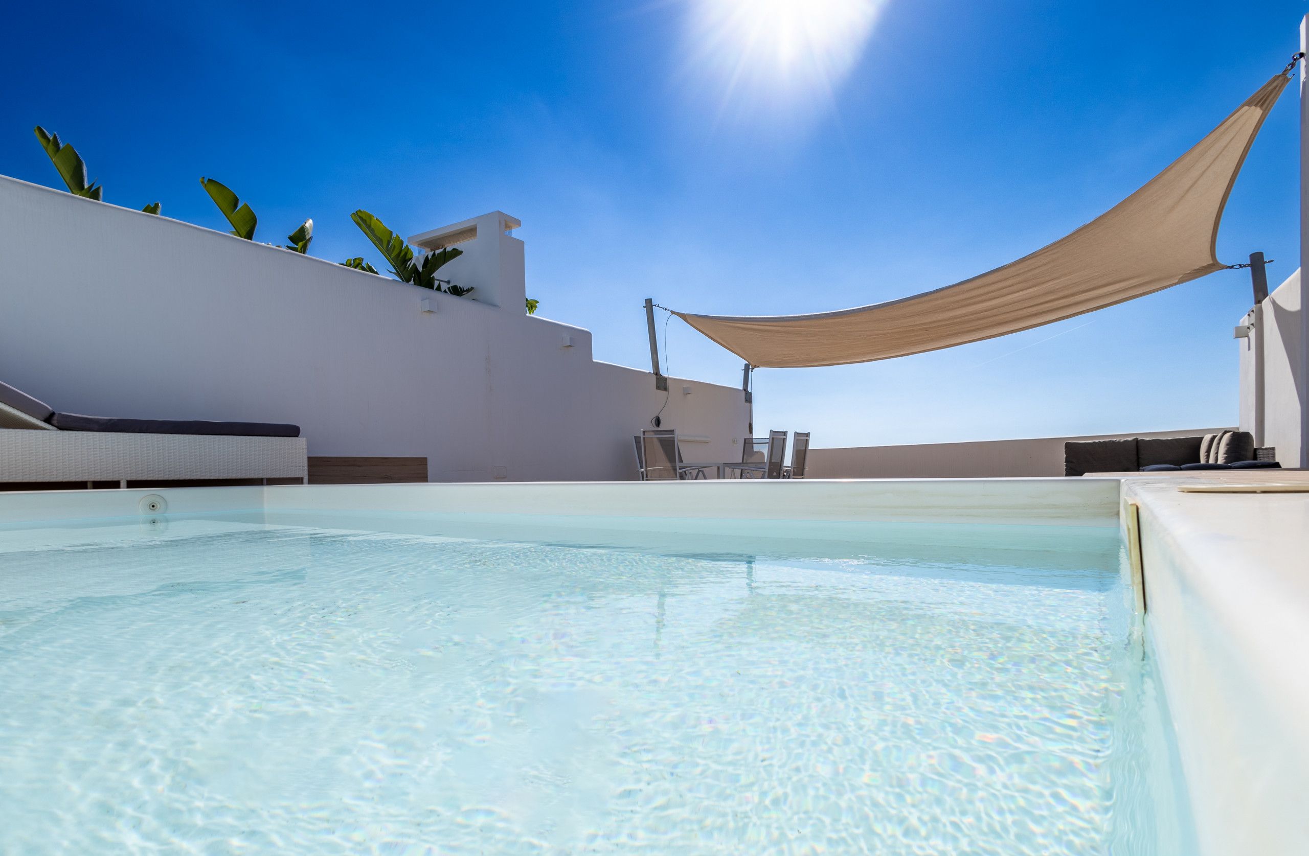 A clear blue pool with lounge chairs and a canopy, surrounded by a white building and greenery.