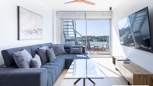 Een moderne woonkamer met een grijze hoekbank, een glazen salontafel, een houten bijzettafeltje, een grote flatscreen-tv en een plafondventilator.
