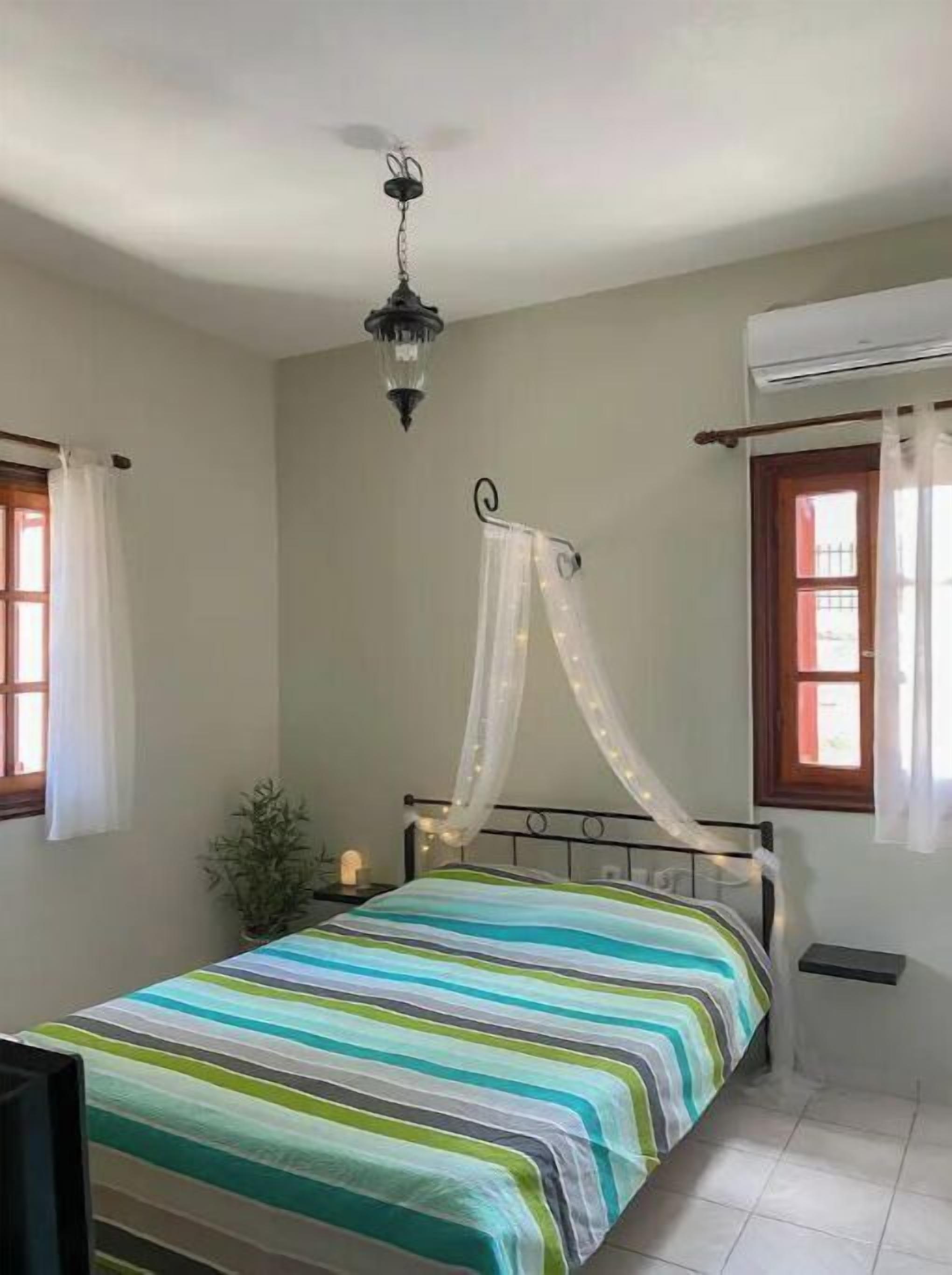 Une chambre à coucher comprenant un lit, un luminaire suspendu, une fenêtre avec des rideaux et un appareil de climatisation.