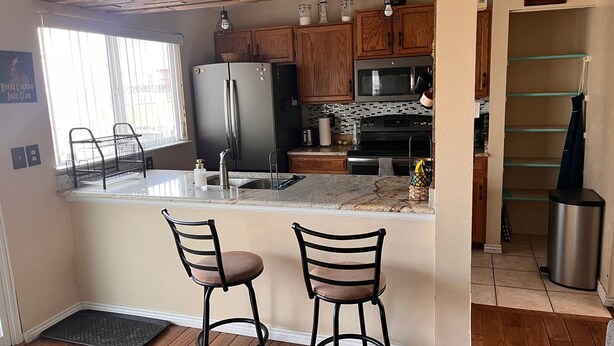 A kitchen with wooden cabinets, a stainless steel refrigerator, a microwave, and a sink. There are two bar stools and a window with blinds. - Spacious home with Tranquil backyard (Aurora)