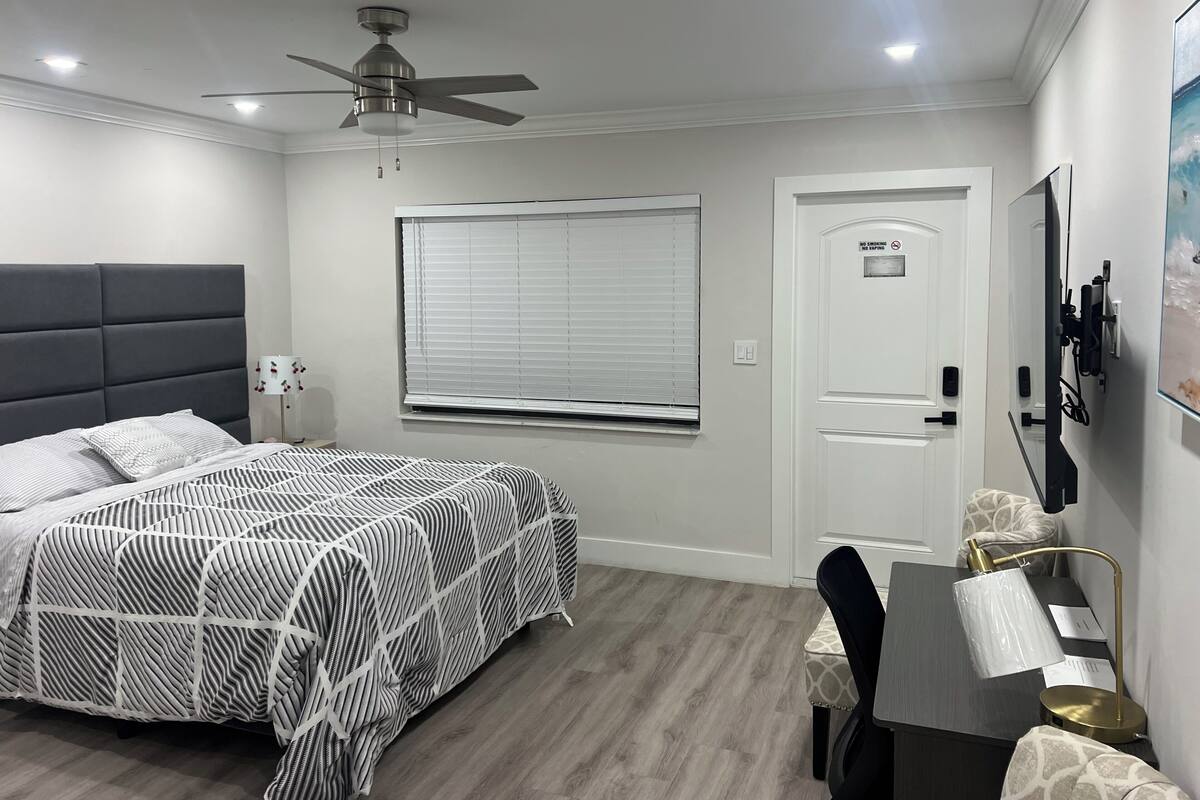 Une chambre à coucher comprenant un lit, un bureau, une chaise, un ventilateur de plafond, une fenêtre avec des stores, une porte et un téléviseur fixé au mur.