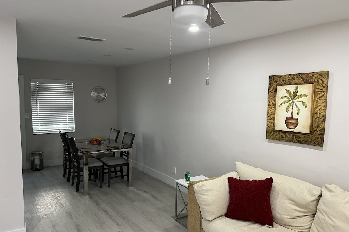 Un salon avec un canapé beige, une table à manger avec des chaises, un ventilateur de plafond et une horloge au mur.