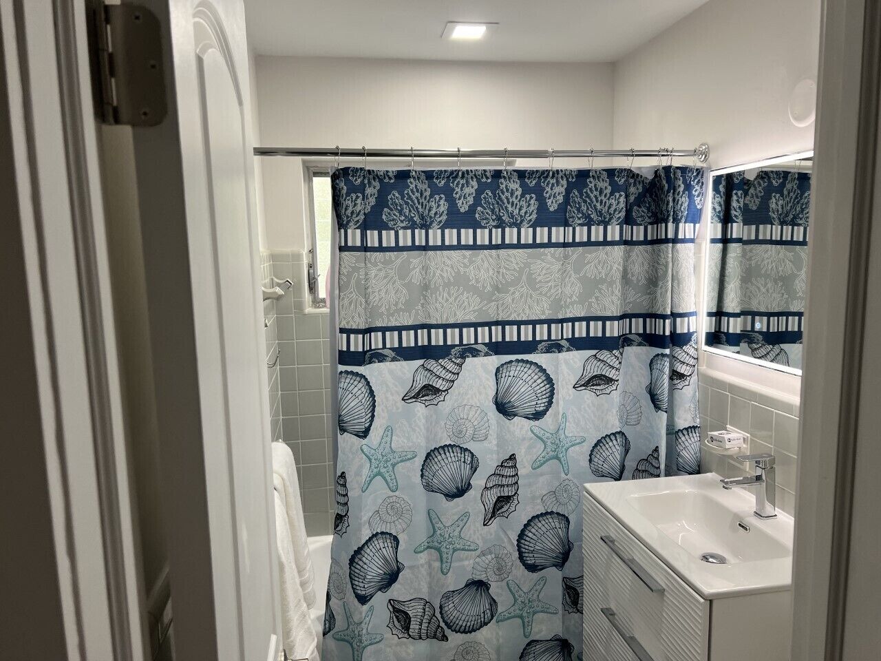 Un baño con cortina de ducha estampada con conchas marinas y estrellas de mar, un lavabo blanco y un espejo encima.