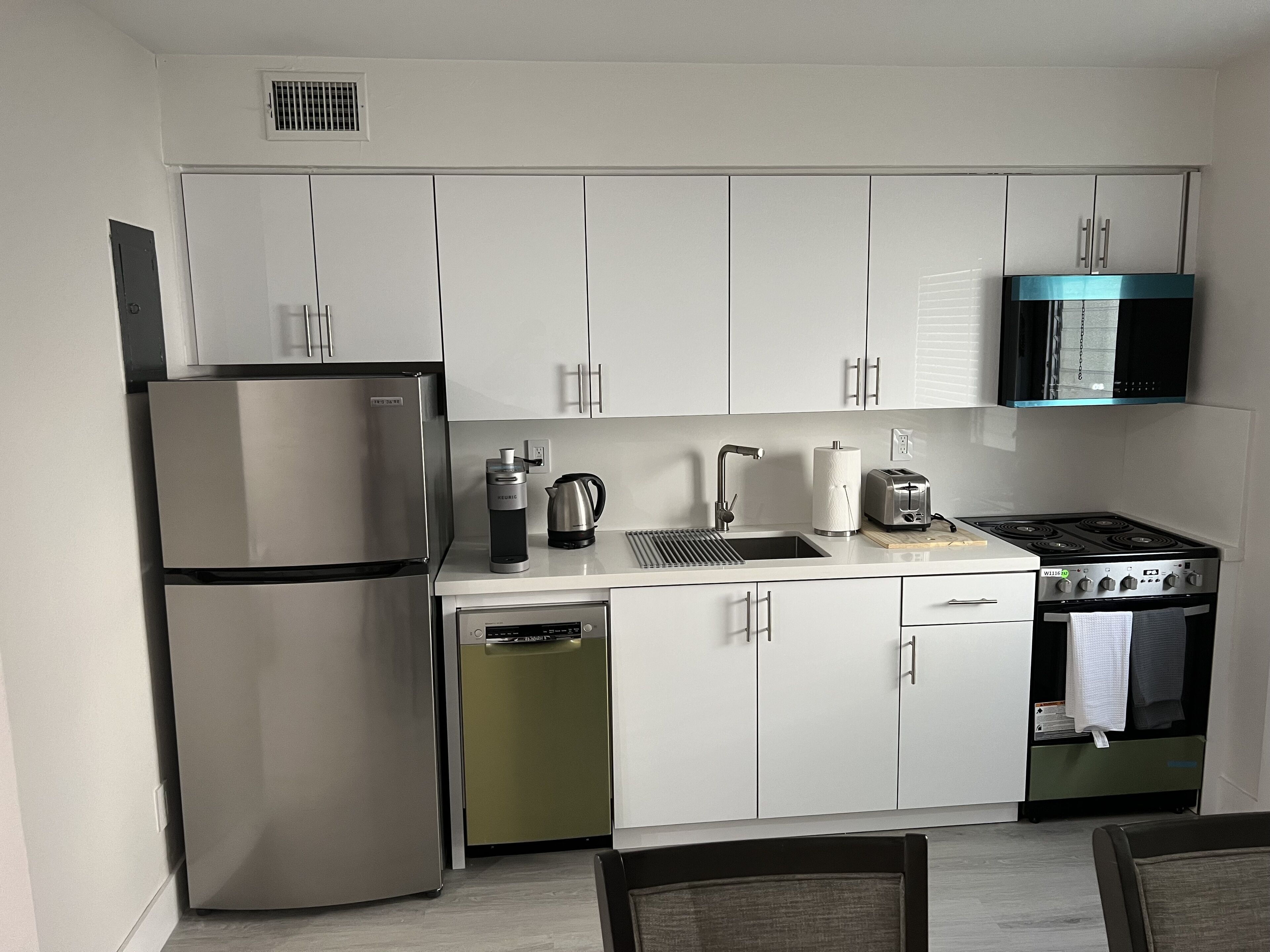 A compact kitchen with white cabinets, a stainless steel refrigerator, a coffee maker, a kettle, a toaster, and a stove.