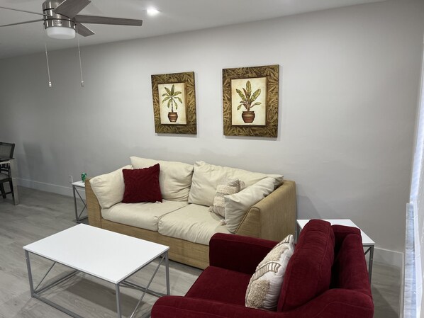 A modern living room with a beige sofa, a red armchair, a white coffee table, and two framed botanical prints on the wall.